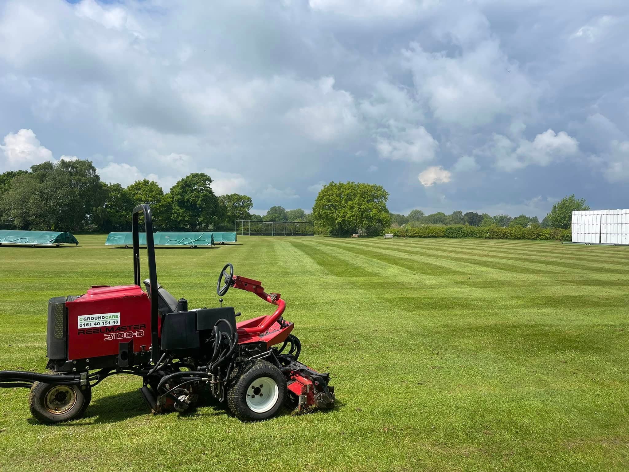 Triple ride-on mower cutting a cricket outfield with a sightscreen behind